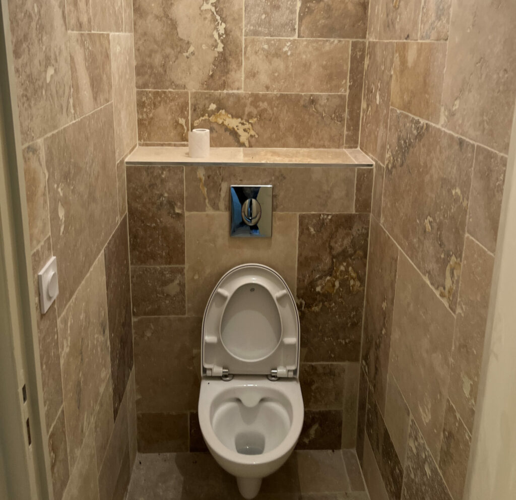 WC suspendu avec plaque de commande chromée, habillage mural en travertin opus et réservation haute pour la future bouche de VMC.