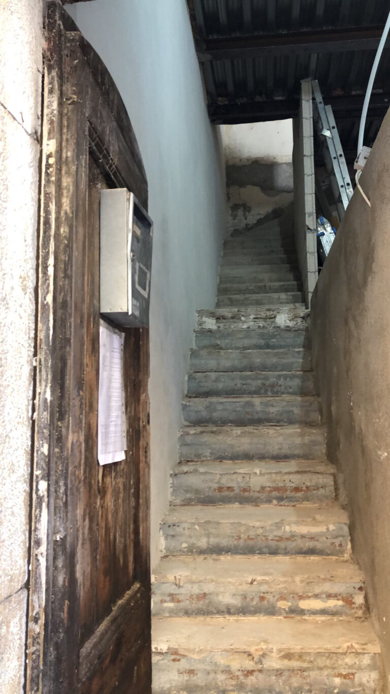 Escalier en béton brut et dégradé dans un couloir étroit, murs non finis et ancienne porte en bois à l’entrée.