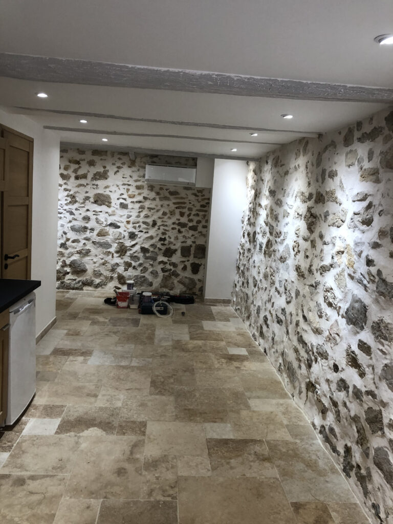 Vue longitudinale de la cave réaménagée en pièce habitable, avec éclairage LED encastré, murs en pierre et dallage en travertin.