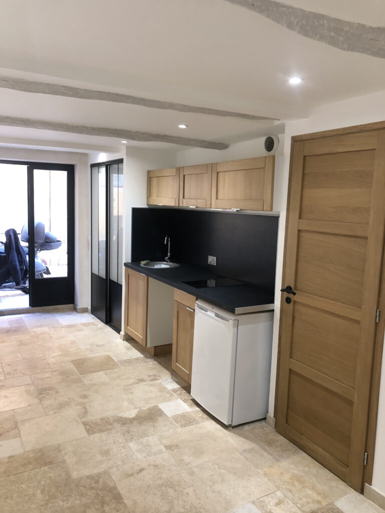 Vue de la kitchenette installée dans la cave rénovée, avec meubles en chêne, plan noir, crédence pleine hauteur et verrière coulissante donnant sur la baie vitrée.