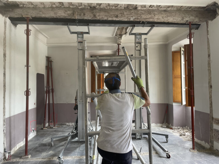 Gros plan sur deux IPN levés à hauteur sous plafond, bridés autour d’une vieille poutre bois, avec étais rouges et deux lève-matériaux en place.