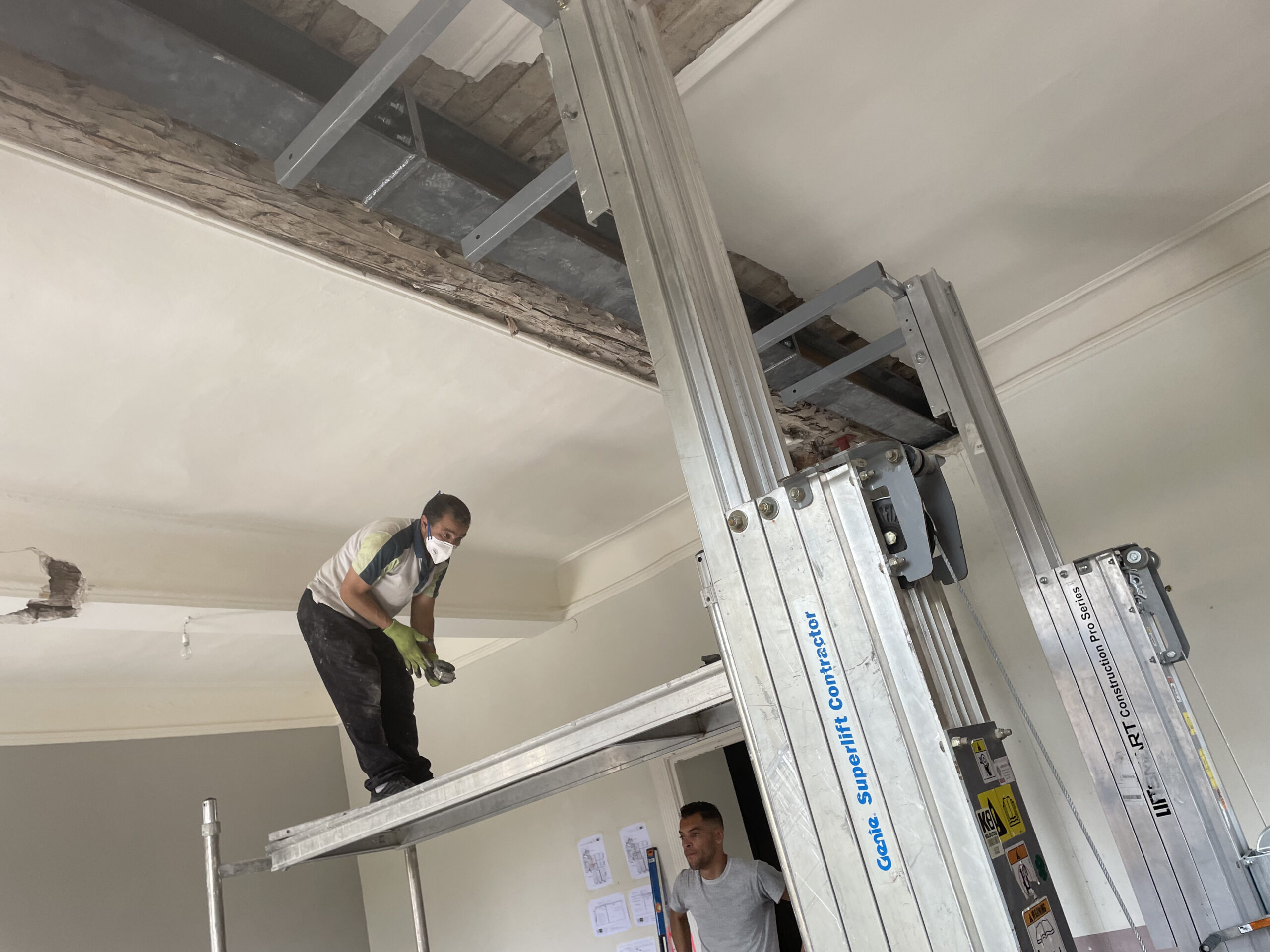 Vue oblique montrant les lève-matériaux Genie soulevant un cadre acier ; fenêtres ouvertes, échafaudage et réservations dans les murs porteurs.