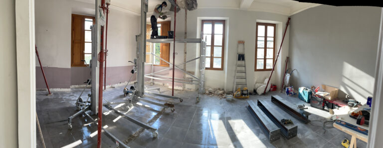 Panorama du chantier : grandes ouvertures, files d’étais, tracés au sol et poutres IPN prêtes à être posées sur le côté.